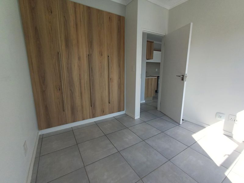 A bright, empty bedroom featuring modern wood-finish built-in cupboards and neutral grey floor tiles, with a doorway leading to an adjacent kitchen area.