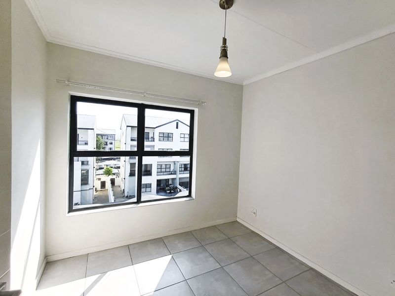 A bright, empty bedroom featuring neutral tiled flooring, a modern pendant light, and a large window with black aluminium frames.