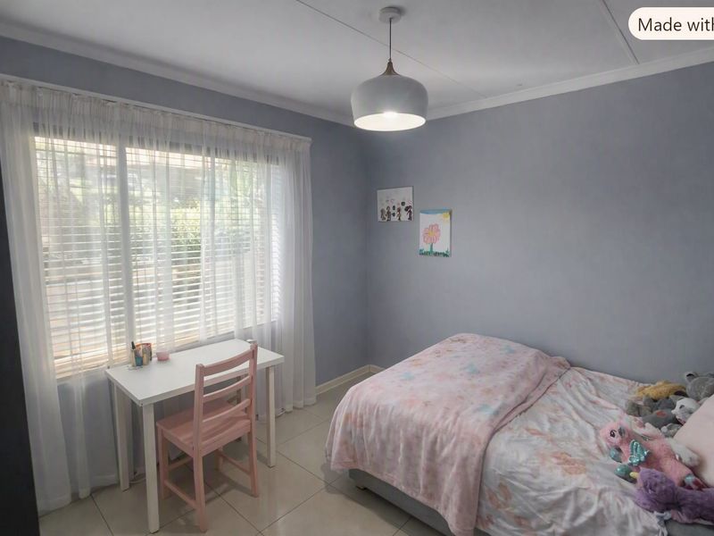 A well-maintained bedroom featuring neutral grey walls, tiled flooring, and large windows fitted with venetian blinds and sheer curtains.