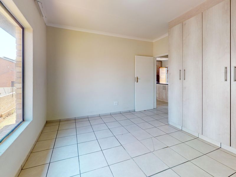 A bright, empty bedroom featuring neutral tiled flooring, built-in cupboards, and a large window with security bars, offering a clean and versatile living space.
