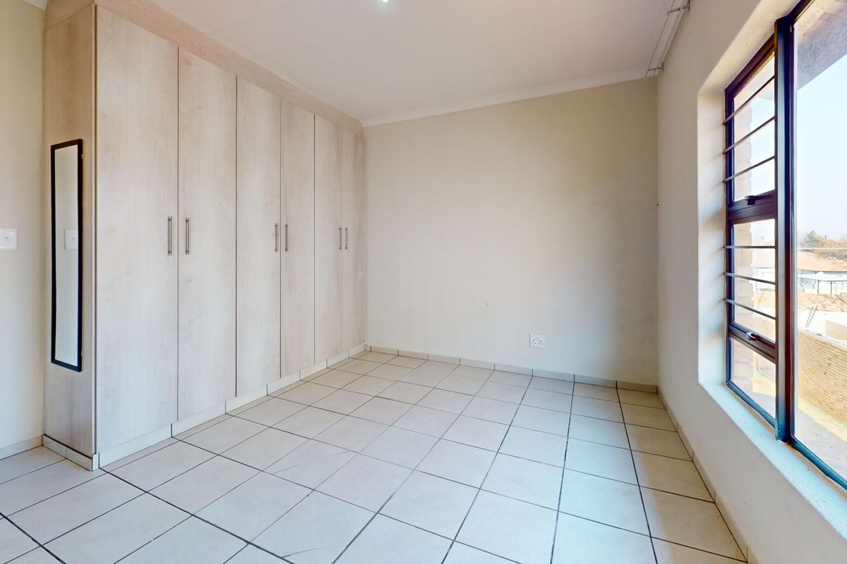 A bright, empty bedroom featuring neutral tiled flooring and ample built-in cupboard storage, complemented by a large window with security bars.