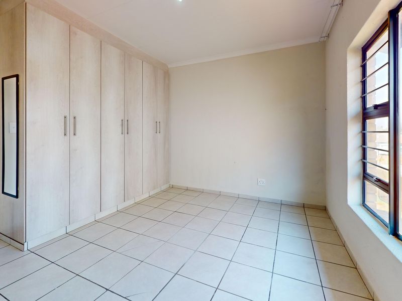 A bright, empty bedroom featuring neutral tiled flooring and ample built-in cupboard storage, complemented by a large window with security bars.