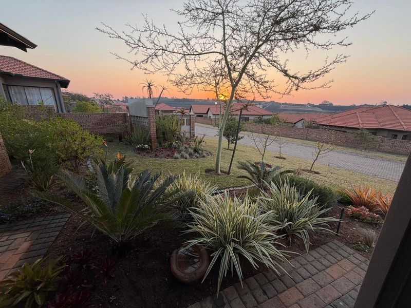 A well-maintained front garden featuring established landscaping, brick paving, and a brick boundary wall, set against a scenic sunset backdrop.