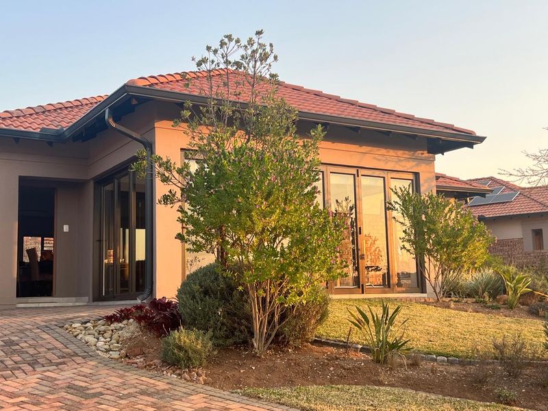 A well-maintained modern house exterior featuring terracotta roof tiles, dark aluminium framed sliding doors, and a neat brick-paved driveway with landscaped garden beds.