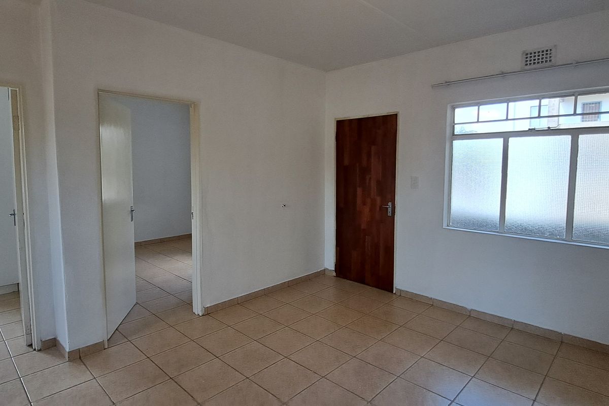 An empty, neutrally painted living area featuring tiled flooring, a timber door, and natural light through an aluminium-framed window.