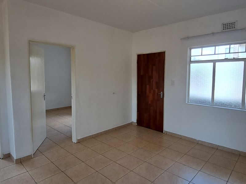 An empty, neutrally painted living area featuring tiled flooring, a timber door, and natural light through an aluminium-framed window.