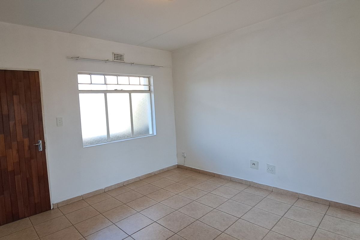 A bright, empty bedroom featuring neutral tiled flooring, a solid timber door, and a window with a ventilation grille.