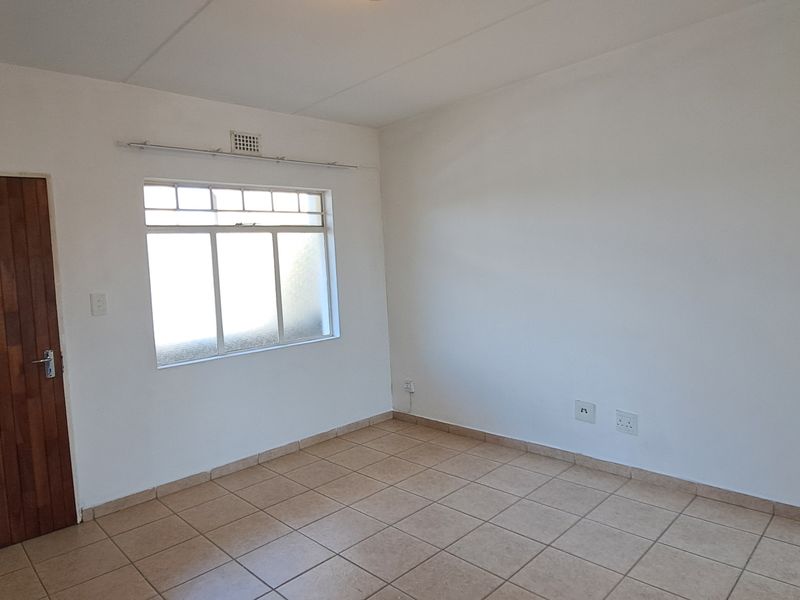 A bright, empty bedroom featuring neutral tiled flooring, a solid timber door, and a window with a ventilation grille.