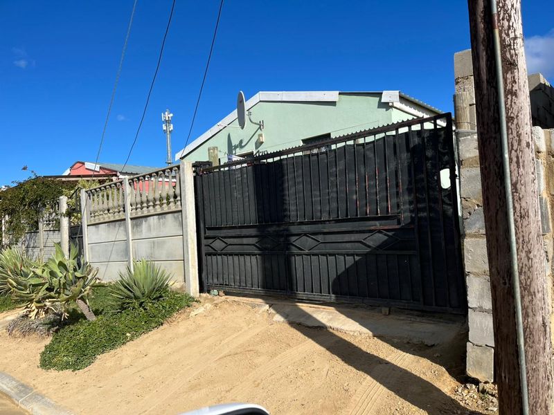 Front view of the property featuring a black metal sliding gate, precast concrete boundary wall, and an unpaved driveway entrance.