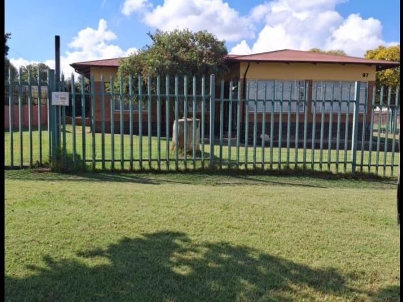 Front view of a single-story brick house featuring a tiled roof and secure metal perimeter fencing set within a grassy yard.