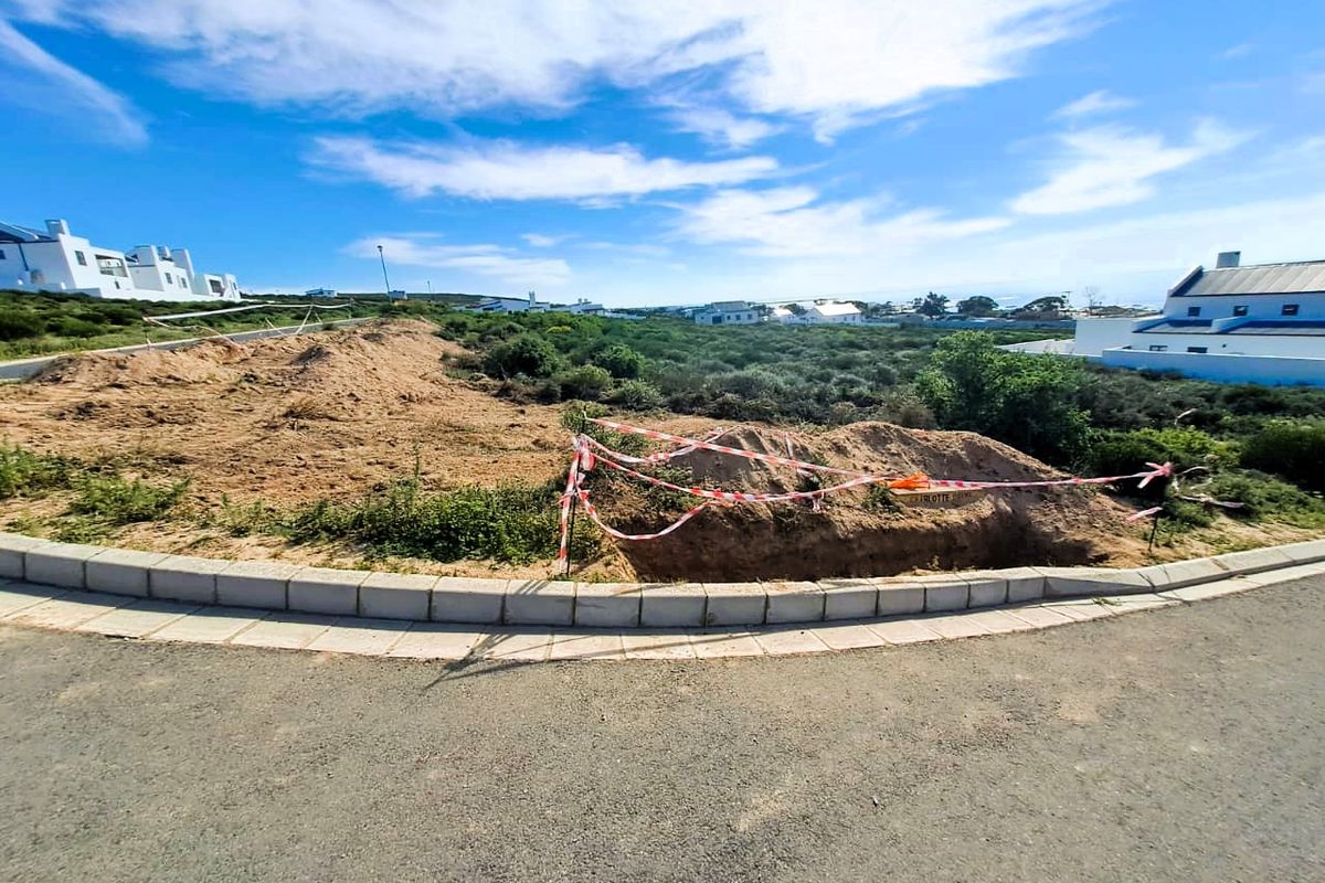 A vacant residential plot featuring an excavated soil mound and construction safety tape, bordered by a paved curb and asphalt road.