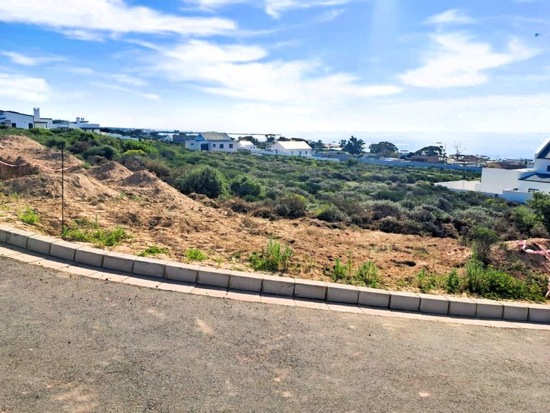 A vacant residential plot featuring concrete kerbing and asphalt road frontage, offering an open landscape with coastal views in the distance.