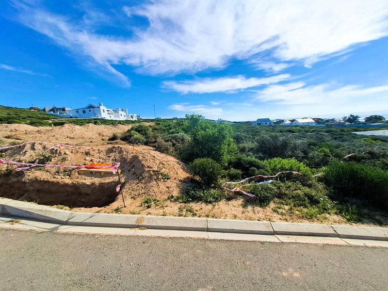 A vacant residential plot featuring an excavated area, bordered by a concrete curb and asphalt road in a coastal neighborhood setting.