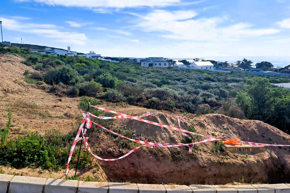 A vacant residential plot featuring an excavated area with safety tape, situated in a coastal neighborhood with surrounding vegetation and distant housing.