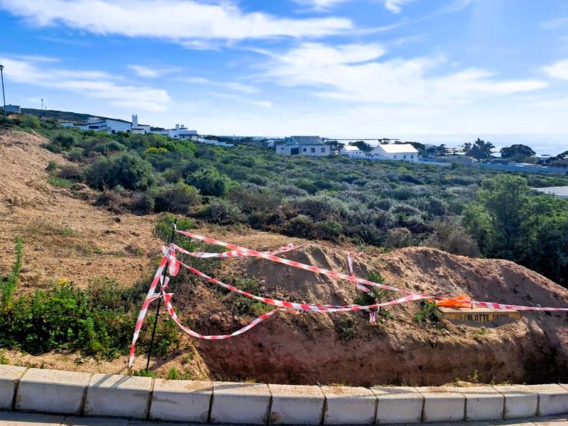 A vacant residential plot featuring an excavated area with safety tape, situated in a coastal neighborhood with surrounding vegetation and distant housing.
