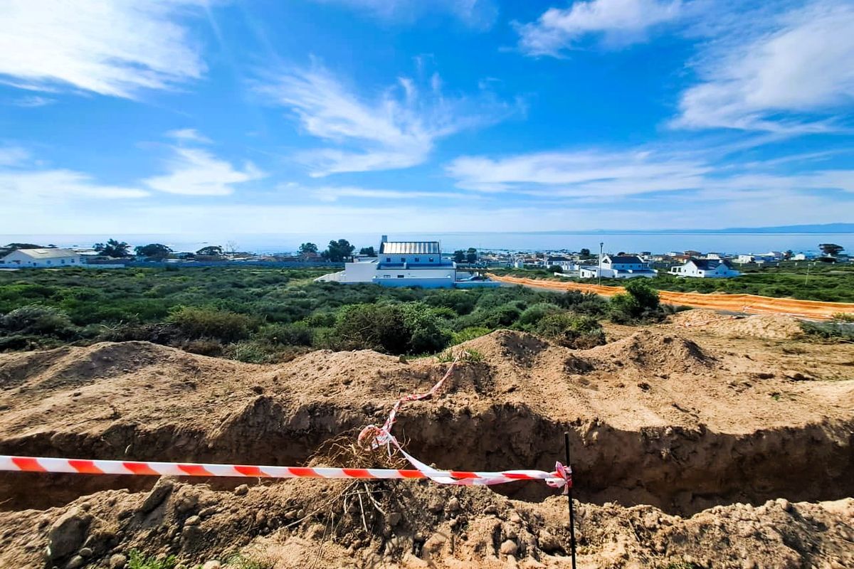 A view of a vacant residential plot featuring an excavated trench and safety tape, with surrounding coastal landscape and distant residential development.