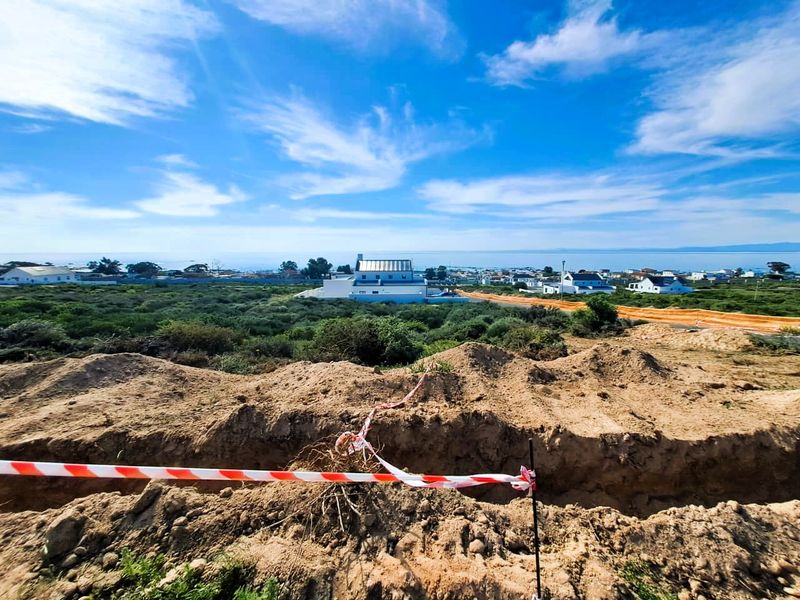 A view of a vacant residential plot featuring an excavated trench and safety tape, with surrounding coastal landscape and distant residential development.