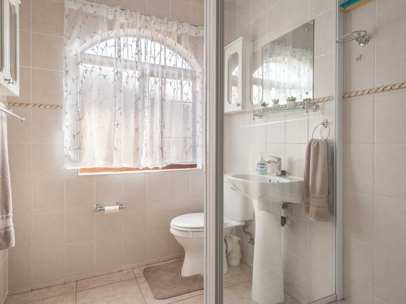 A functional bathroom featuring a pedestal basin, toilet, and a glass-enclosed shower, finished with neutral wall and floor tiles and an arched window.