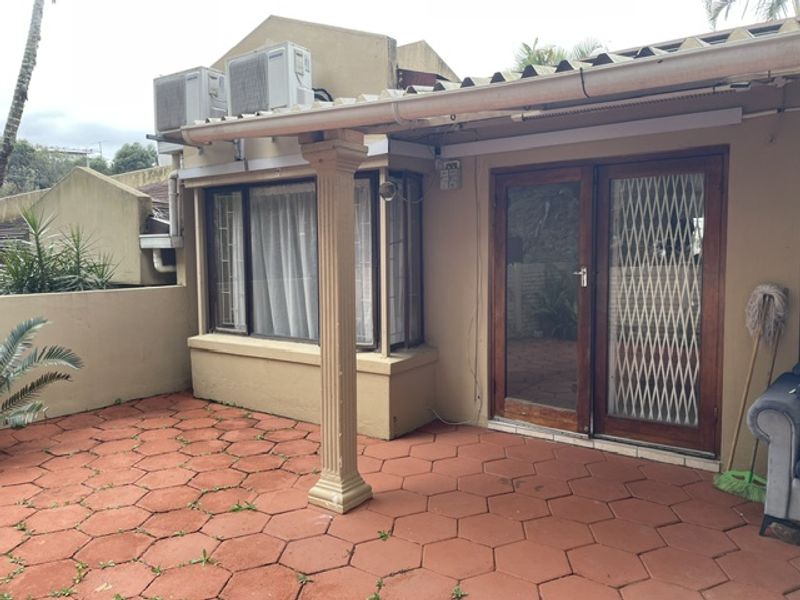 A covered patio area featuring paved flooring, a bay window, and external air conditioning units, providing a functional outdoor space for the apartment.