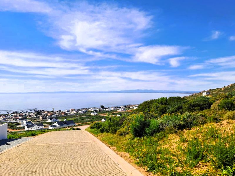 A scenic elevated view overlooking the coastal landscape, residential area, and ocean, featuring a paved driveway in the foreground.