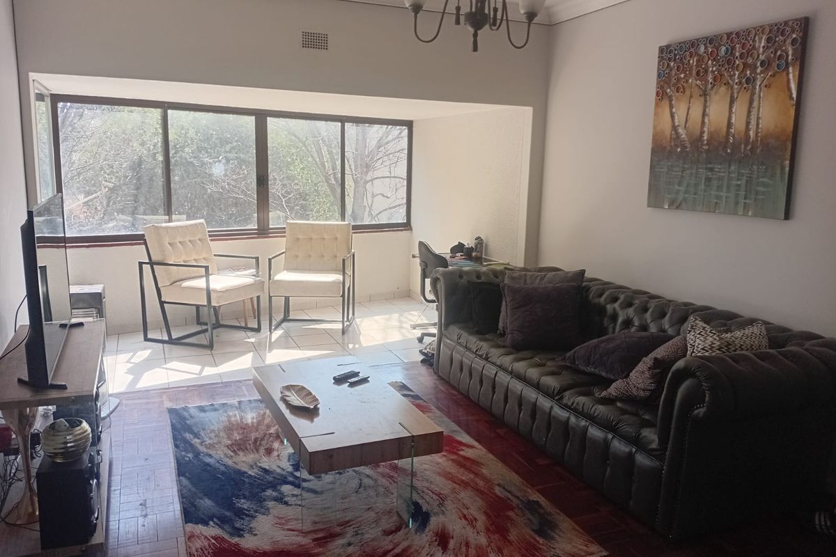 A spacious living room featuring a mix of parquet and tiled flooring, large windows allowing natural light, and classic ceiling cornicing.