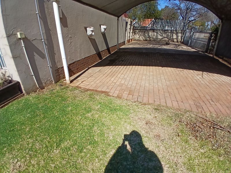 A paved carport area with a covered structure, adjacent to a lawn and boundary wall, showing signs of wear on the exterior wall finish.