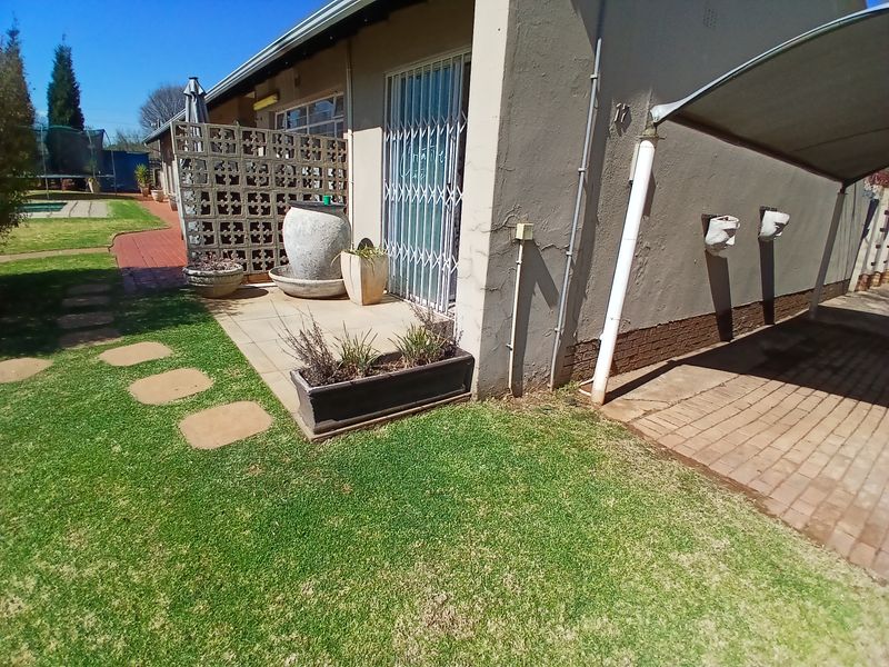 Rear exterior view showing a paved patio area, security gate, carport structure, and landscaped garden space.