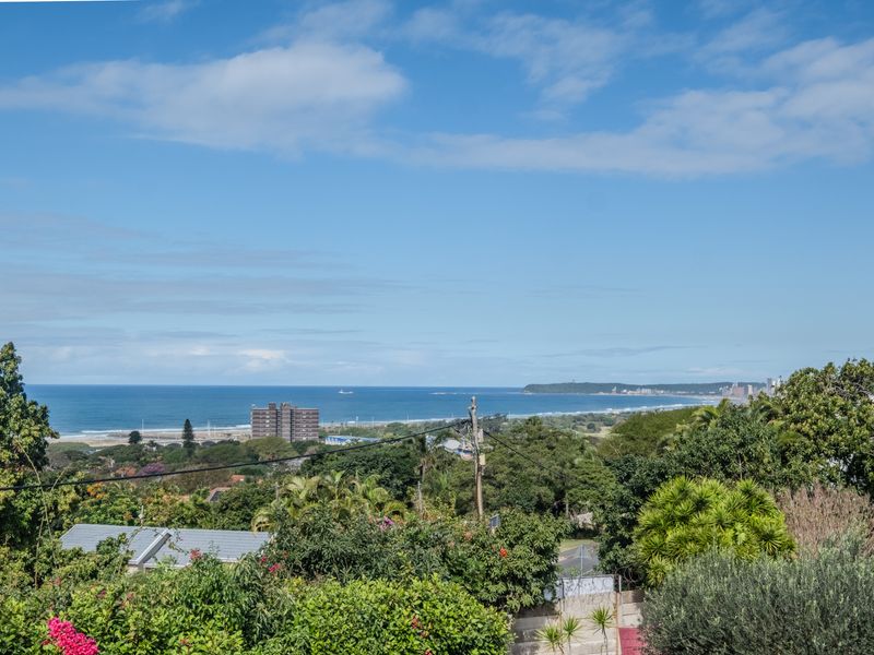 A scenic elevated view overlooking the coastline, featuring lush greenery, urban development, and the ocean horizon under a clear blue sky.