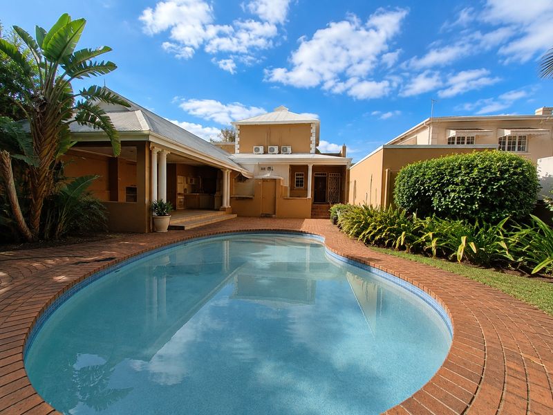 A well-maintained outdoor pool area featuring a swimming pool with brick coping, adjacent to a covered patio with structural columns and exterior air conditioning units.
