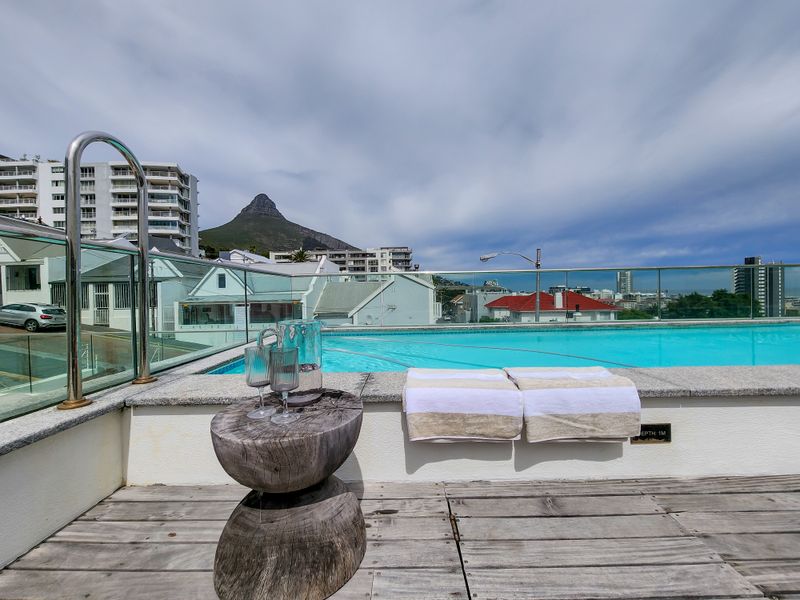 Rooftop swimming pool area featuring a glass balustrade, stone coping, and timber decking with panoramic mountain and city views.