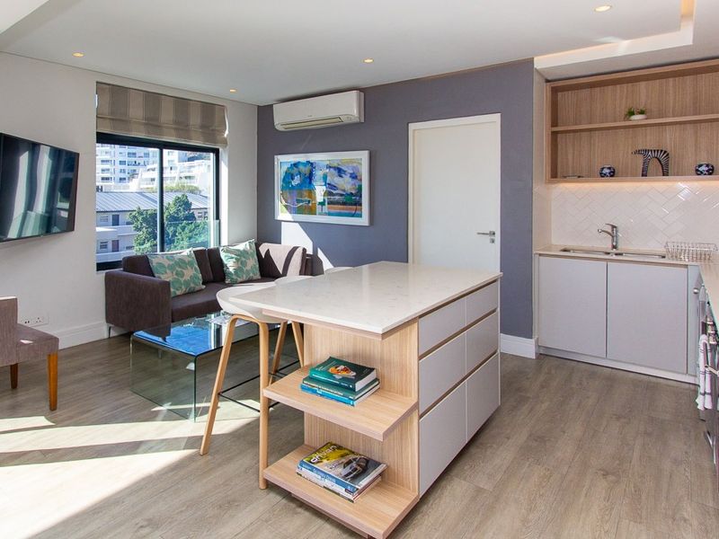 Modern open-plan living area featuring a sleek kitchen with island, herringbone backsplash, and integrated cabinetry, complemented by light wood-look flooring and air conditioning.