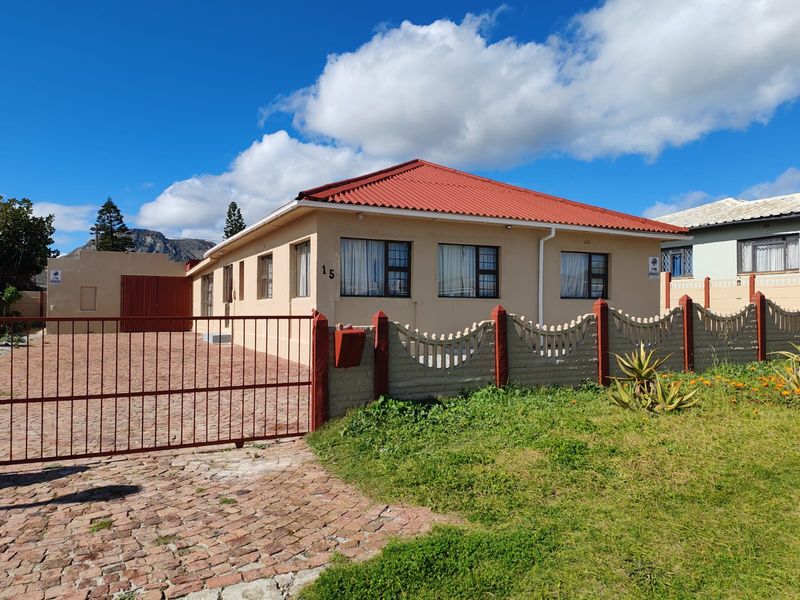 Front view of a single-story house featuring a red corrugated metal roof, paved driveway, and a secure gated entrance with a boundary wall.