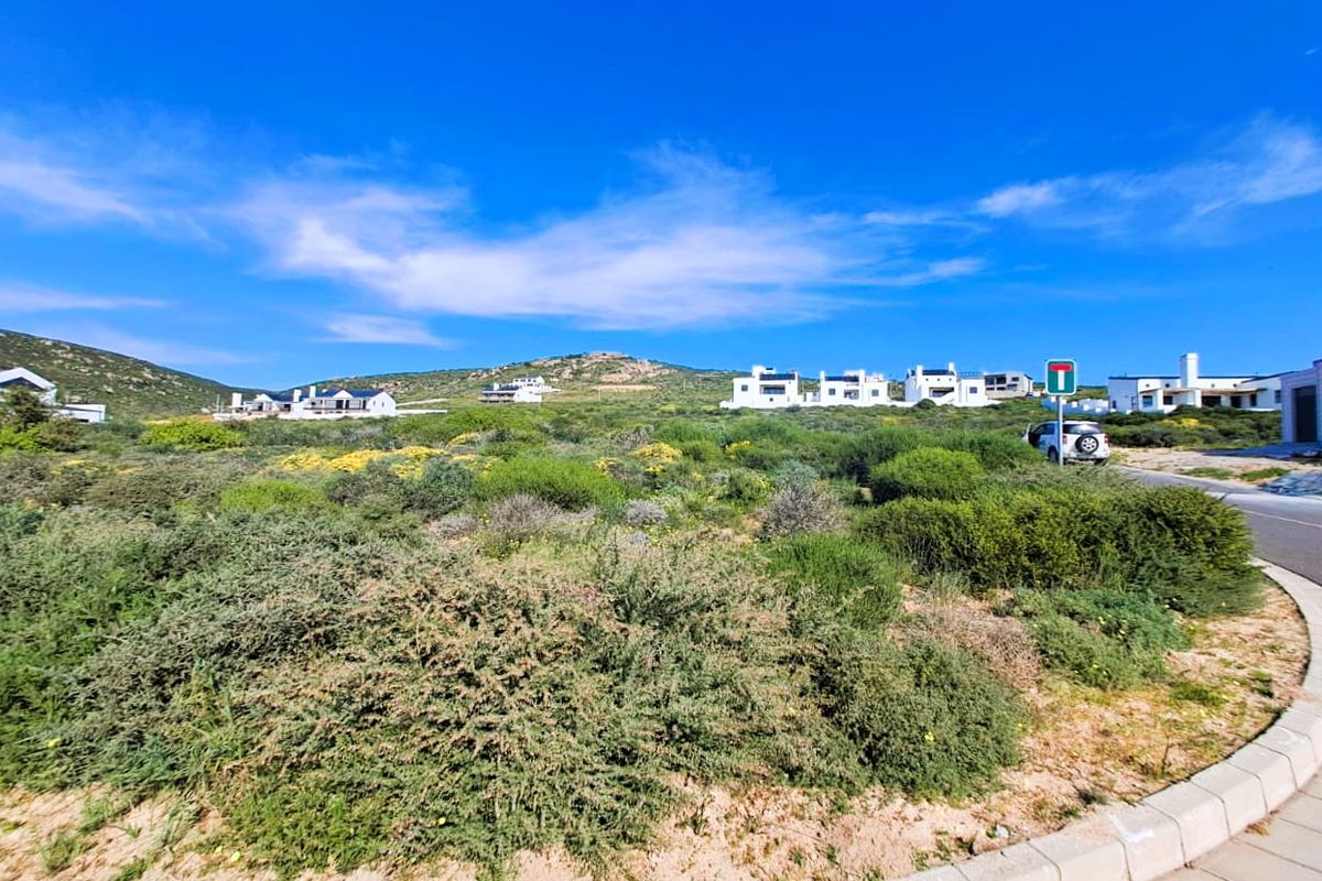 A vacant residential plot featuring natural coastal vegetation, paved curbing, and road access within a developed estate setting.