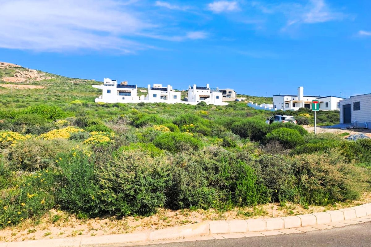 A view of a vacant residential plot featuring natural coastal vegetation, with established residential properties visible in the background.