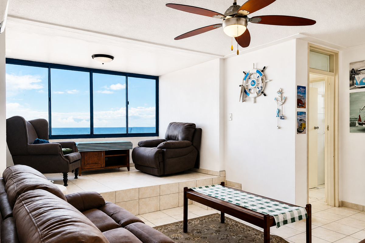 A living room featuring a raised tiled platform, ceiling fan, and large windows offering ocean views, with direct access to an adjacent room.