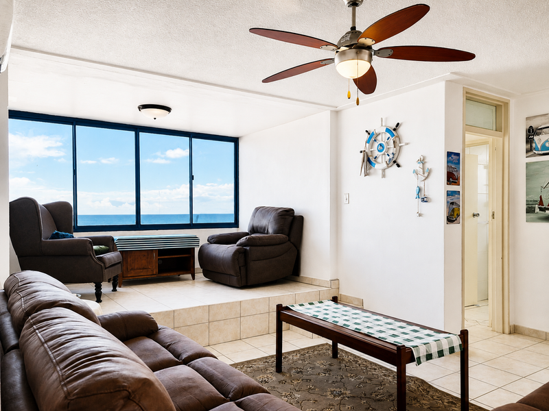 A living room featuring a raised tiled platform, ceiling fan, and large windows offering ocean views, with direct access to an adjacent room.