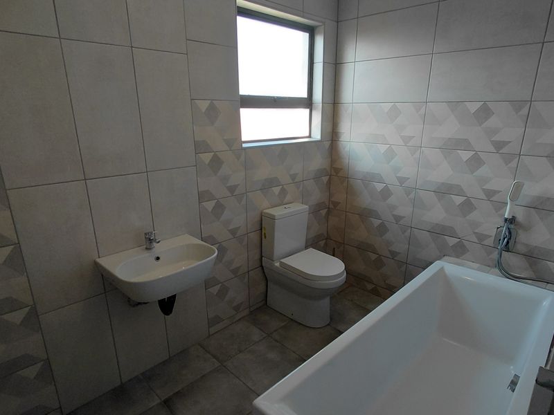 A modern, clean bathroom featuring neutral geometric wall tiles, a wall-mounted basin, a dual-flush toilet, and a white bathtub.