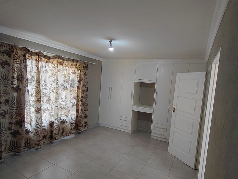 A spacious bedroom featuring neutral tiled flooring, built-in cupboards with an integrated vanity, and large windows with curtain fixtures.