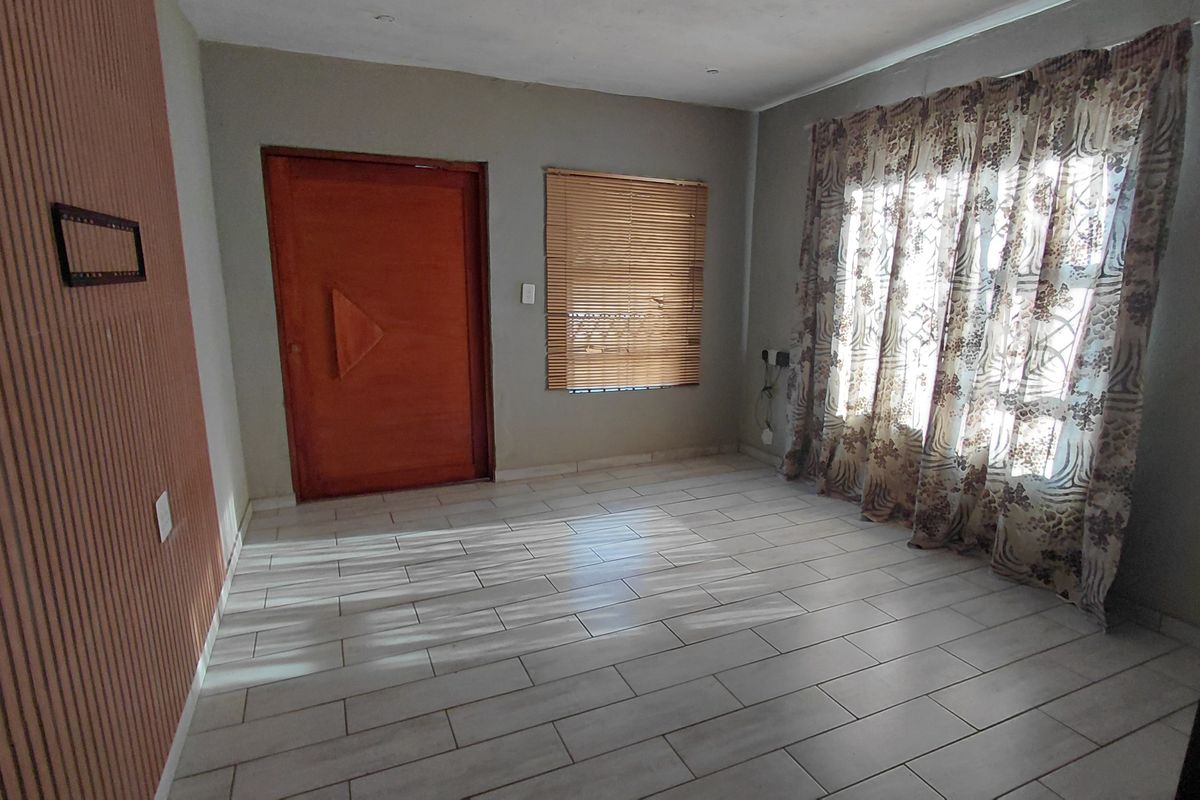 An empty living room featuring light-colored tiled flooring, a wooden entrance door, venetian blinds, and decorative wall paneling.