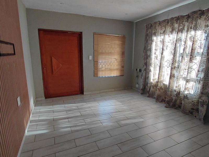 An empty living room featuring light-colored tiled flooring, a wooden entrance door, venetian blinds, and decorative wall paneling.