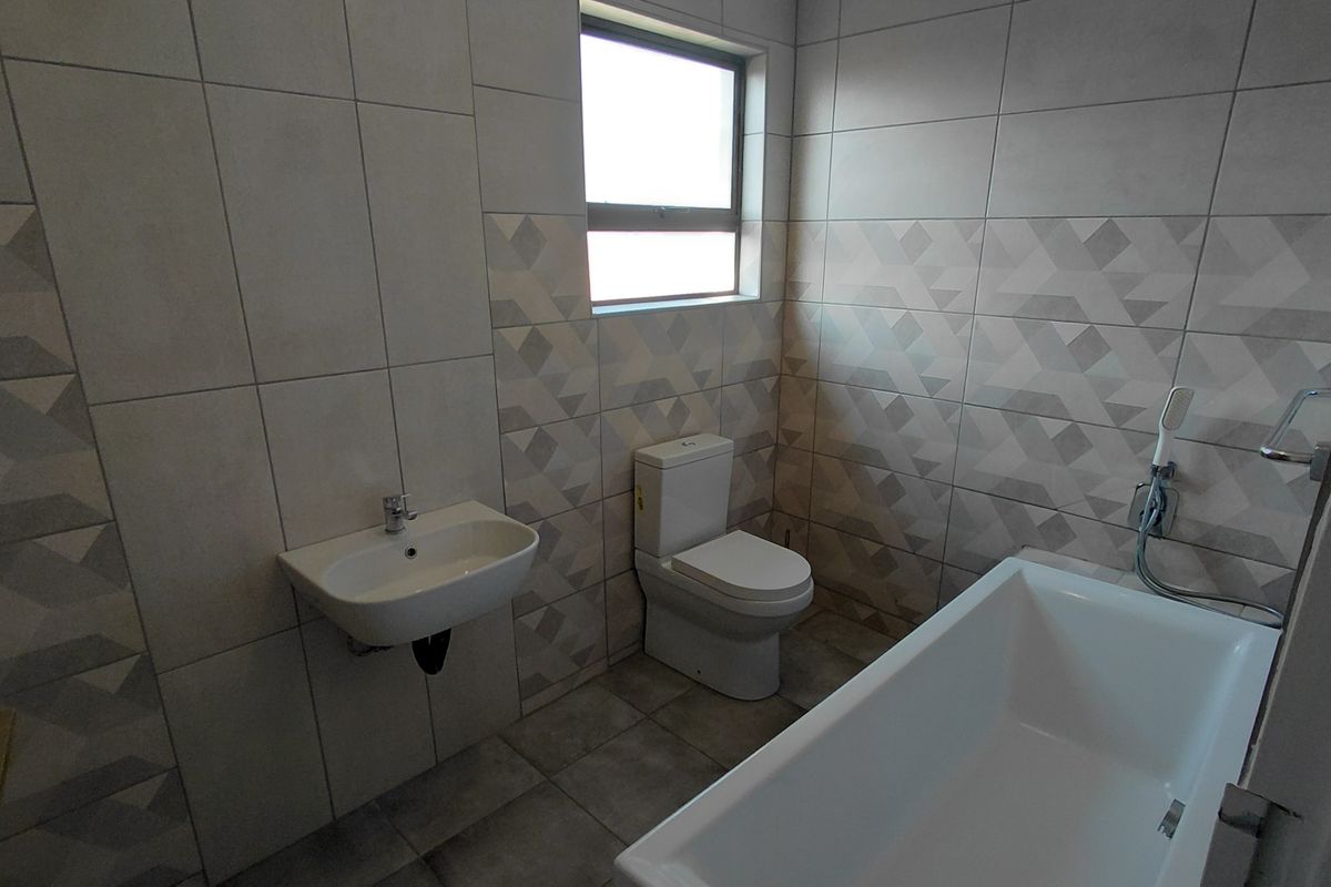 A modern, clean bathroom featuring neutral geometric wall tiles, a wall-mounted basin, a dual-flush toilet, and a white bathtub.