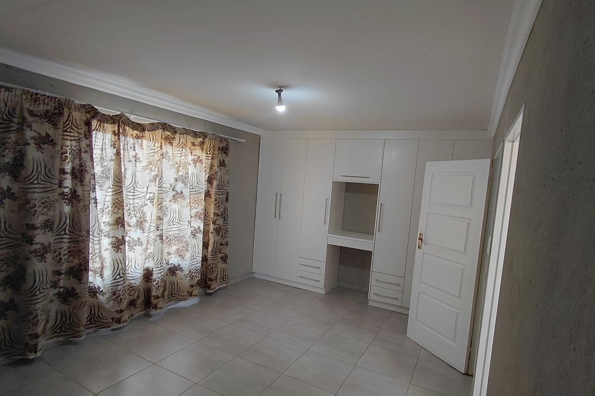 A spacious bedroom featuring neutral tiled flooring, built-in cupboards with an integrated vanity, and large windows with curtain fixtures.