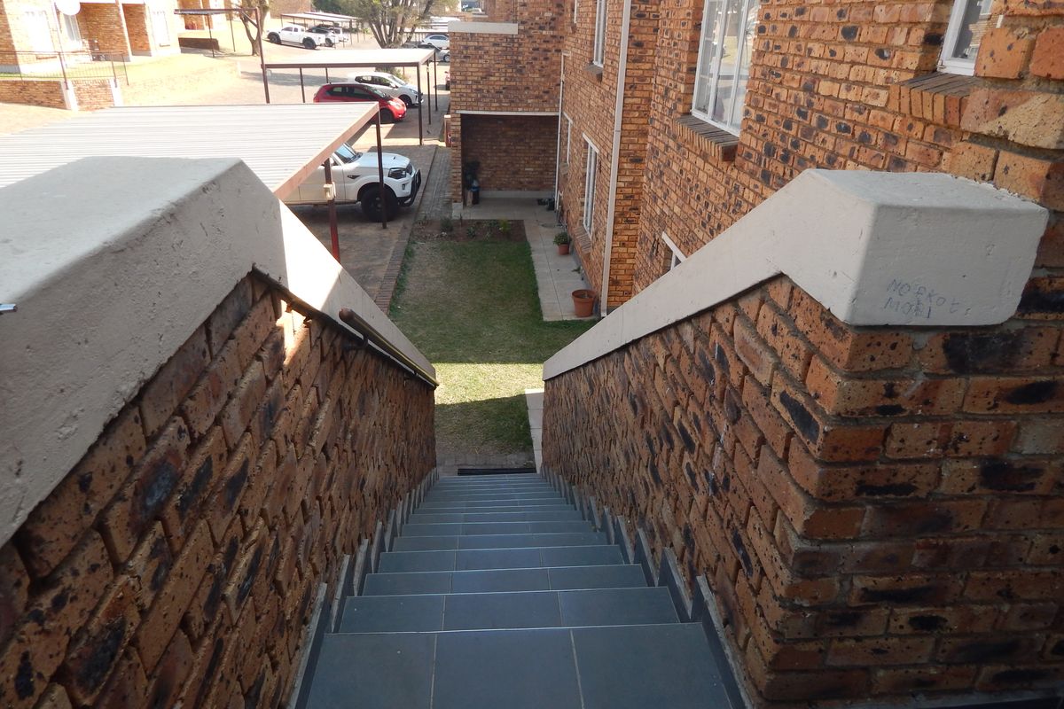An elevated view looking down a brick staircase with tiled steps, leading towards a communal garden and carport area within the townhouse complex.