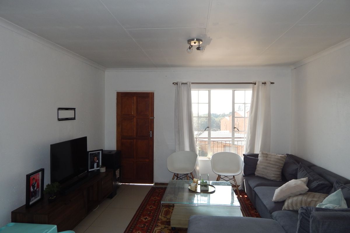 A functional living room featuring tiled flooring, a wooden entrance door, and a window with security bars, offering a bright and practical living space.