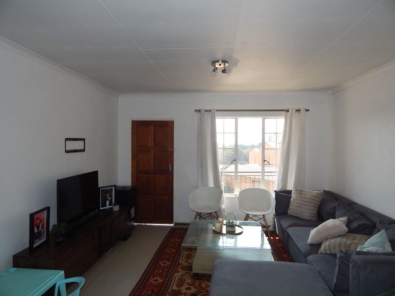 A functional living room featuring tiled flooring, a wooden entrance door, and a window with security bars, offering a bright and practical living space.