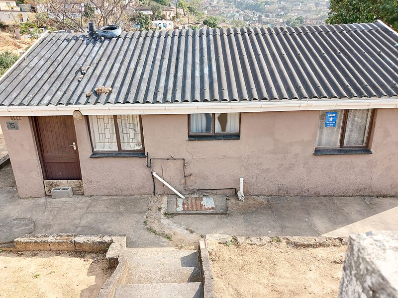 Rear exterior view of a single-story house featuring a corrugated roof, security bars on windows, and concrete stairs leading to the building.