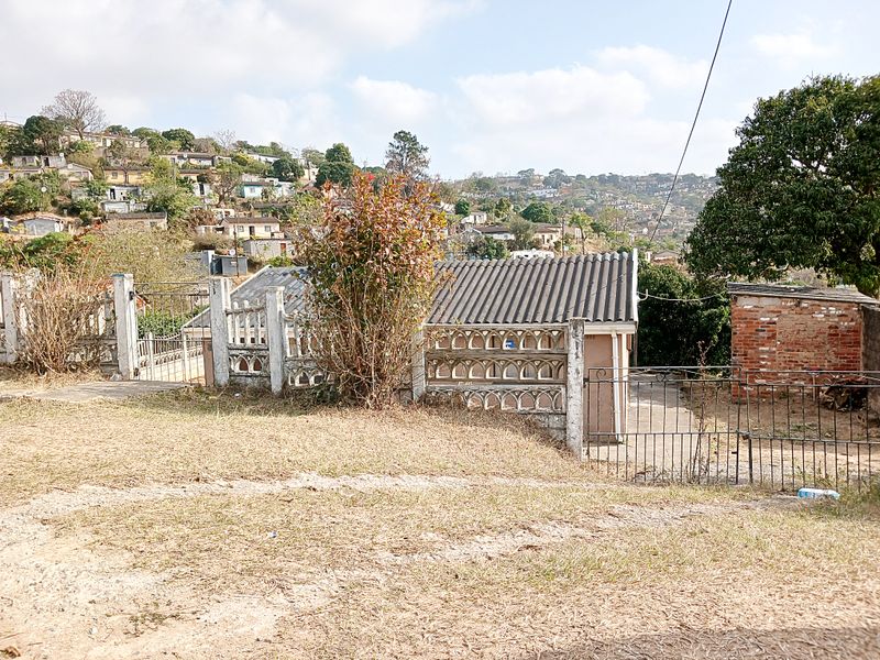 Front view of the property featuring a concrete boundary wall, wrought iron gate, and a small brick outbuilding set against a hillside residential backdrop.