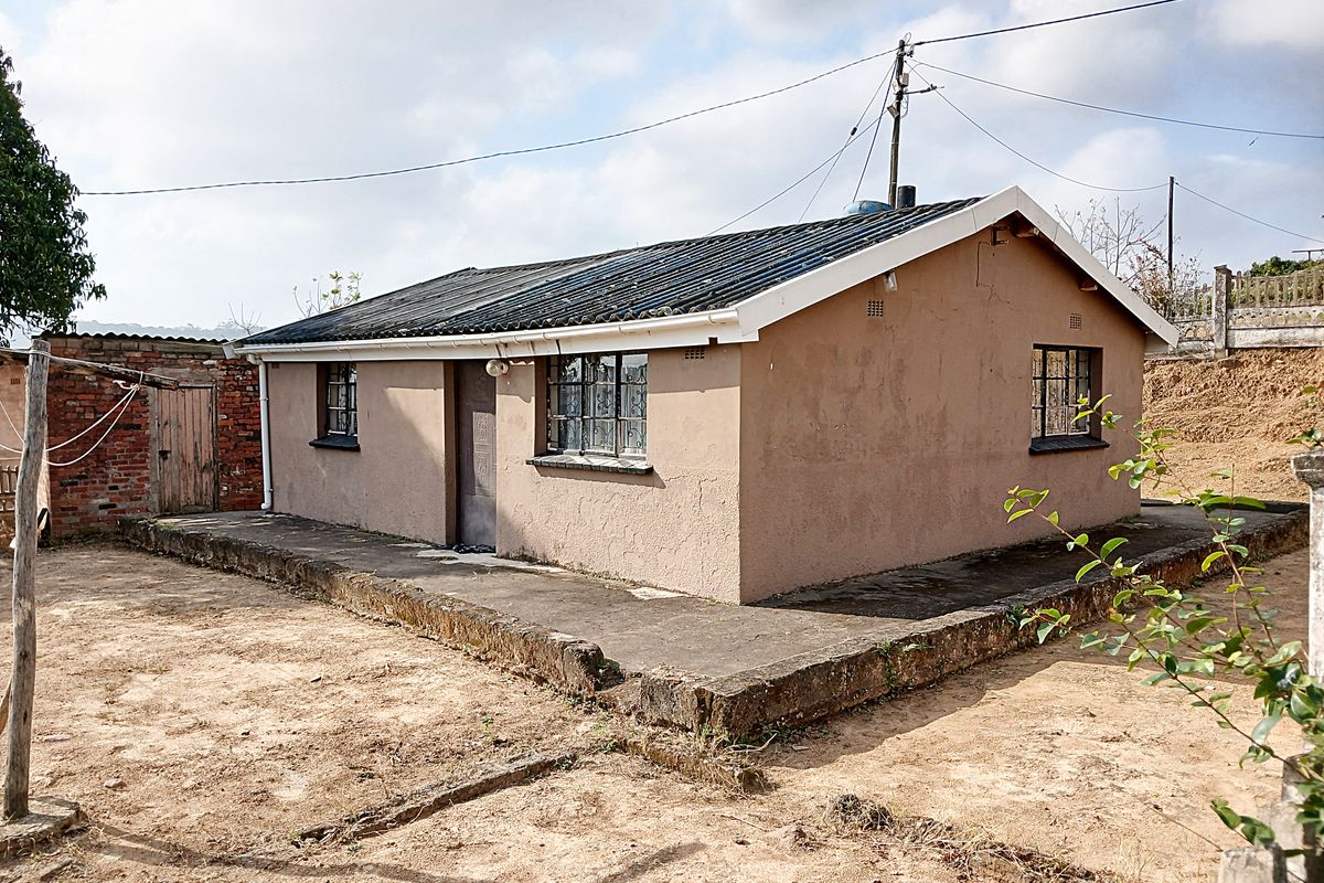 A detached single-story dwelling featuring a corrugated iron roof, steel-framed windows, and a surrounding concrete apron set on a dirt plot.
