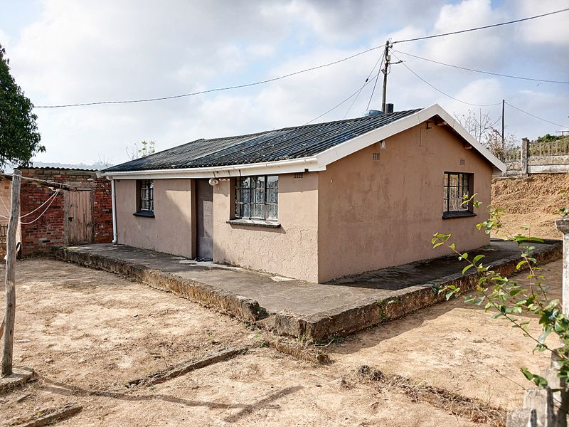 A detached single-story dwelling featuring a corrugated iron roof, steel-framed windows, and a surrounding concrete apron set on a dirt plot.