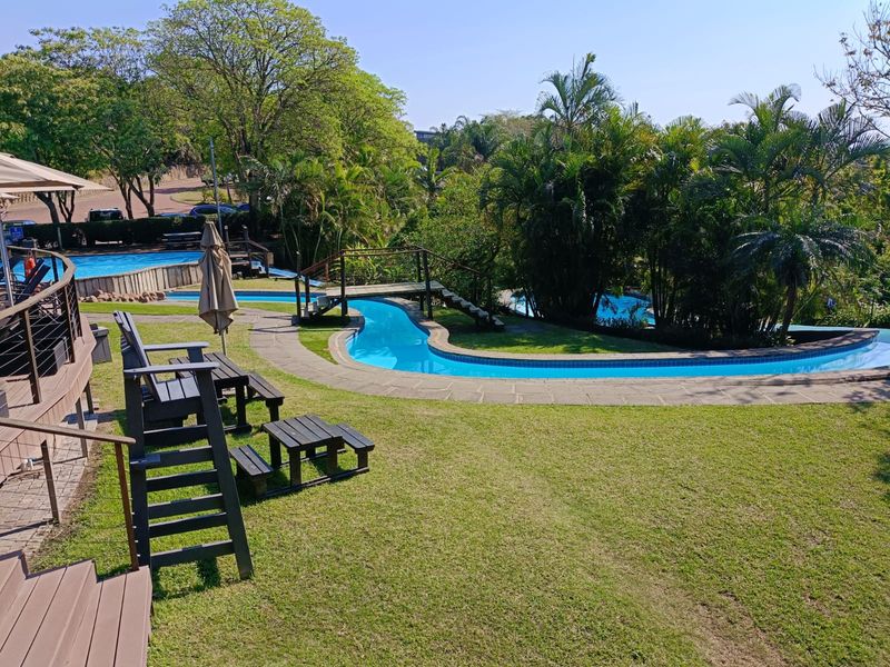 A well-maintained communal pool area featuring a winding swimming pool, wooden footbridge, landscaped garden, and timber decking for outdoor relaxation.